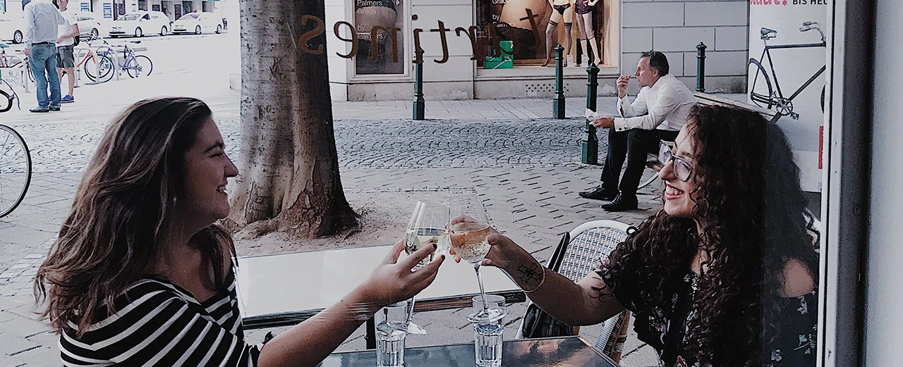 © Jelena Colic Zwei Frauen stoßen mit Weingläsern an einem Straßencafé an.  Freundinnen treffen sich im Freien.