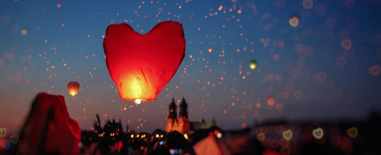 © canva Herzförmige Himmelslaterne mit vielen weiteren Laternen am Abendhimmel über einer Stadt. Romantisches Bild.