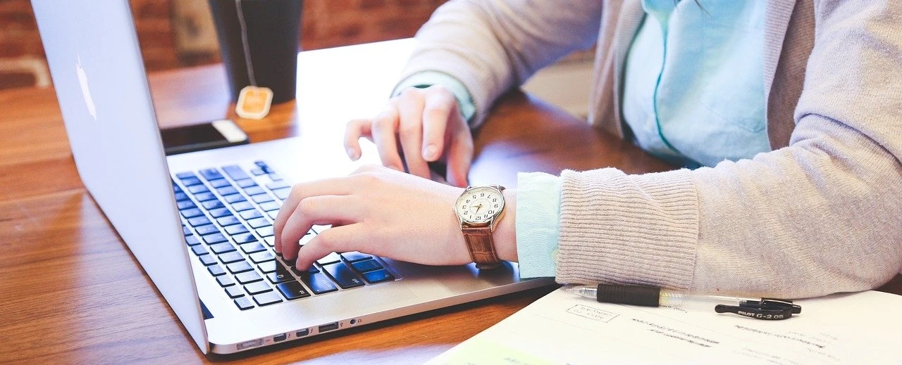 Nahaufnahme: Hände tippen auf Laptoptastatur; Papiere liegen daneben.