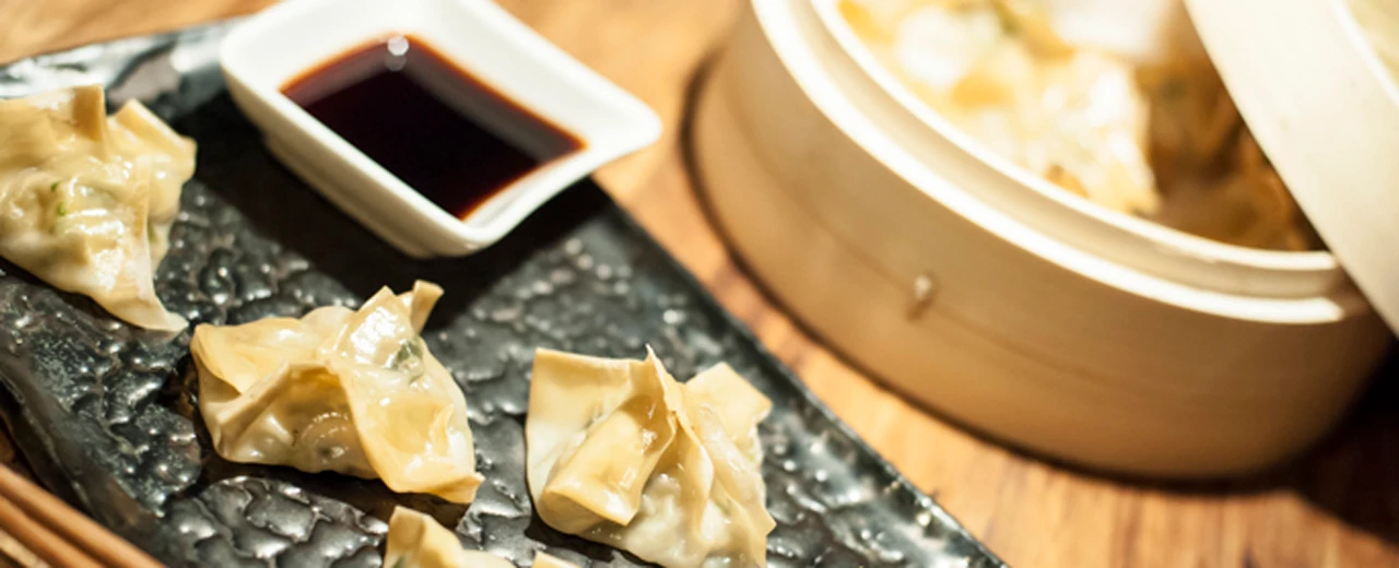 Gedämpfte Teigtaschen mit Sojasauce auf dunklem Teller neben Bambusdampfkorb.