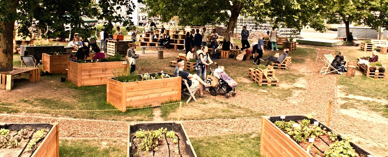 Urbane Gartenlandschaft mit Hochbeeten aus Holz, Palettenmöbeln und Menschen, die sich entspannen. Foto aufgenommen mit einer SONY DSC Kamera.