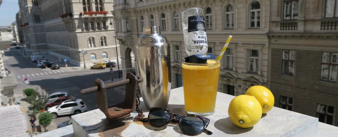 © Anna Gugerell Erfrischendes Corona-Getränk mit Zitronen, Cocktailshaker & Sonnenbrille auf Balkon mit Stadtblick.
