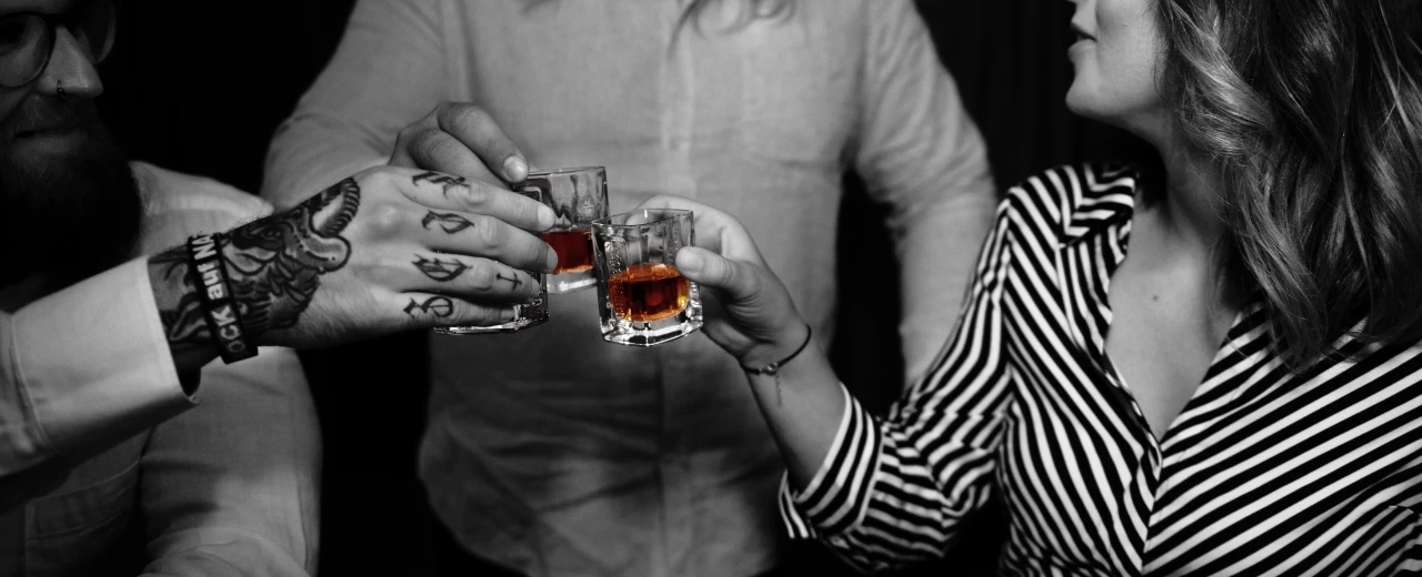 Drei Personen stoßen mit Gläsern mit bernsteinfarbenem Getränk an. Schwarzweiß-Foto mit farbigen Getränken.