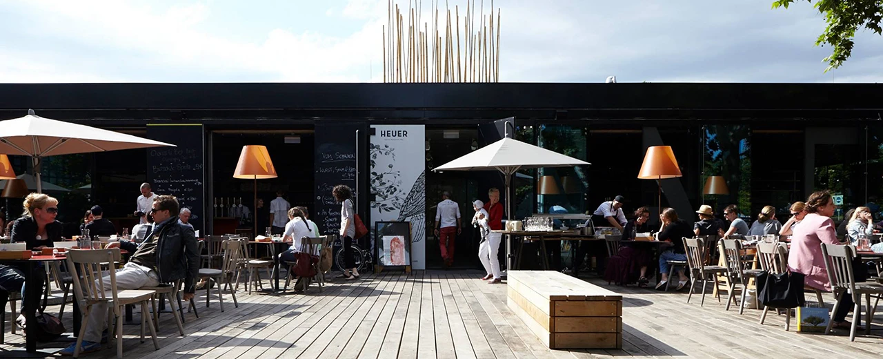 © Heuer am Karlsplatz Sonnige Außenterrasse eines modernen Restaurants mit Gästen an Tischen unter Sonnenschirmen.  Holzterrassenboden, moderne Möbel.