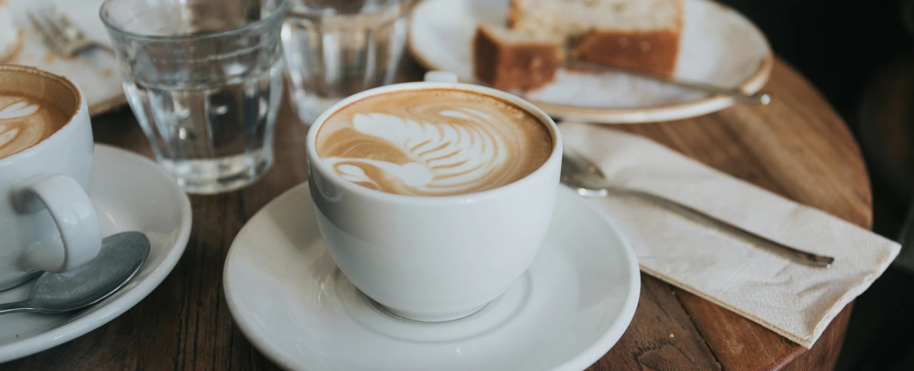 Zwei Tassen Cappuccino mit Latte Art, Wassergläser, Kuchen und Besteck auf Holztisch. Café-Atmosphäre.