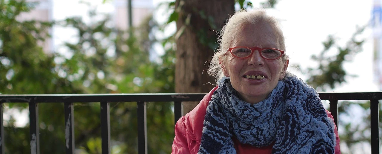 Porträt ältere Frau, blonde Haare, rote Brille, roter Mantel, blauer Schal, Bäume Hintergrund