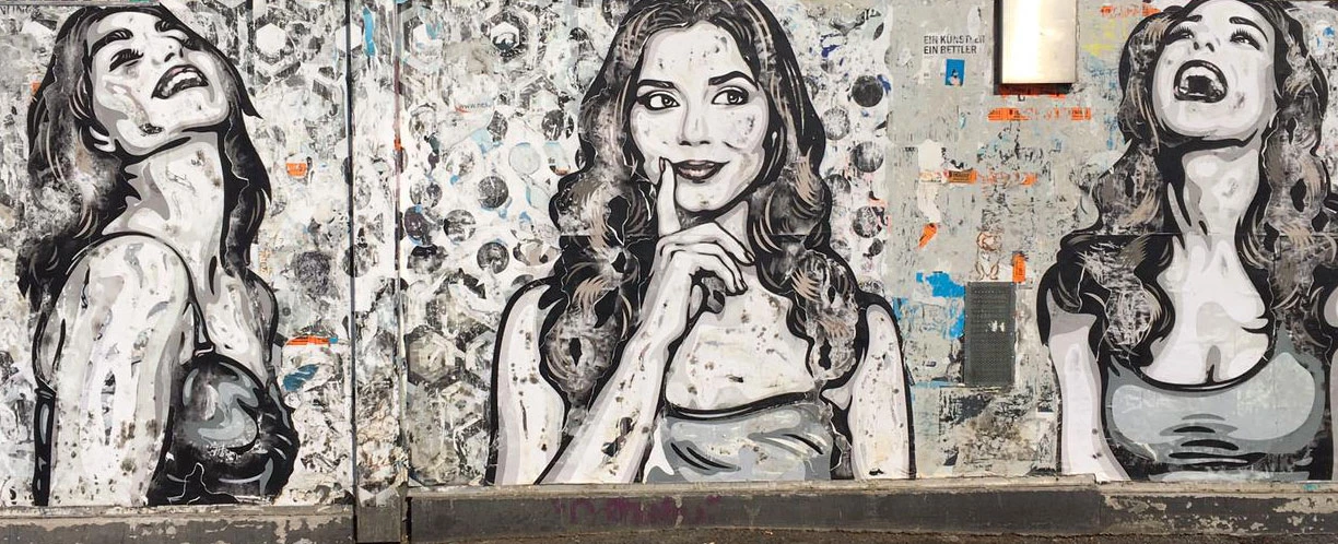 Streetart Wandbild: Drei Frauenporträts in Schwarz-Weiß auf einer mit Plakaten überklebten Wand.