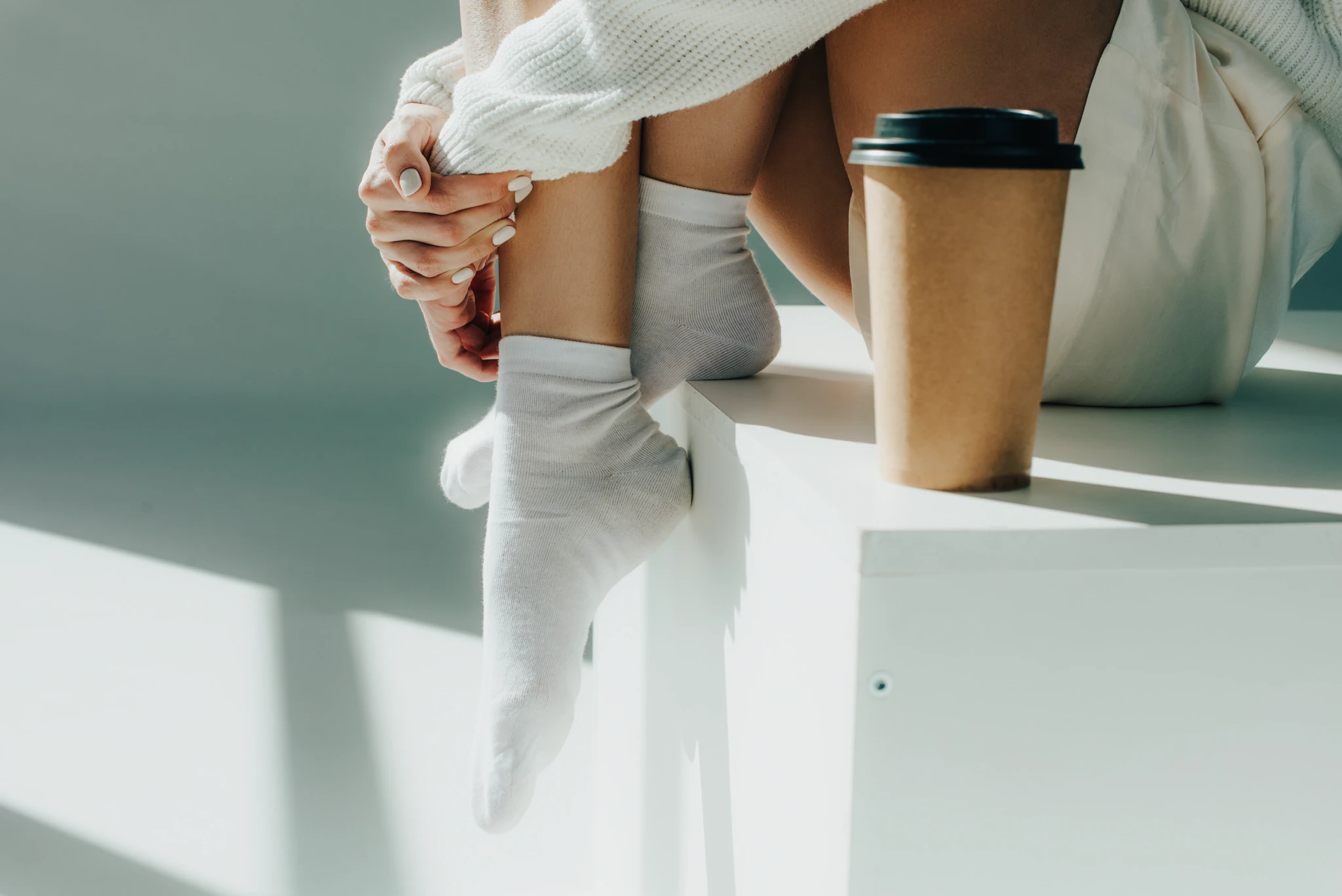 Füße einer jungen Frau, sitzend auf Kasten und neben steht ein Becher Kaffee