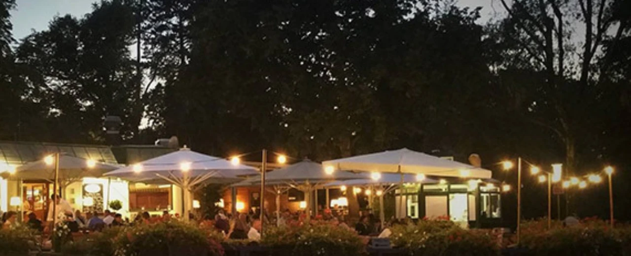 Gemütlicher Restaurant-Außenbereich am Abend, beleuchtet mit Lichterketten und Sonnenschirmen. Gäste sitzen an Tischen.