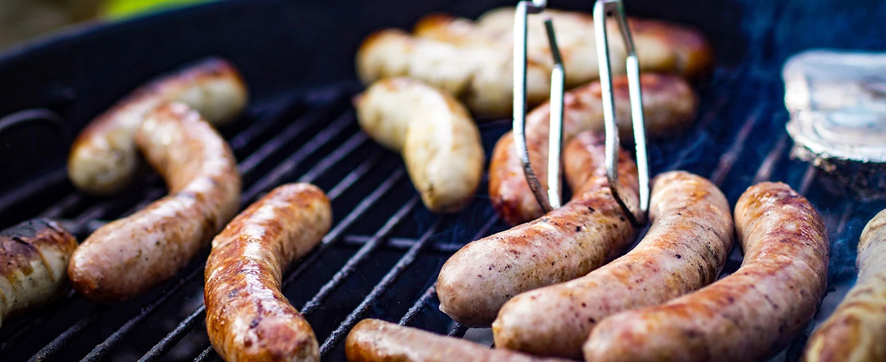 Nahaufnahme von Grillwürsten, die auf einem heißen Grillrost liegen und mit einer Grillzange gewendet werden.